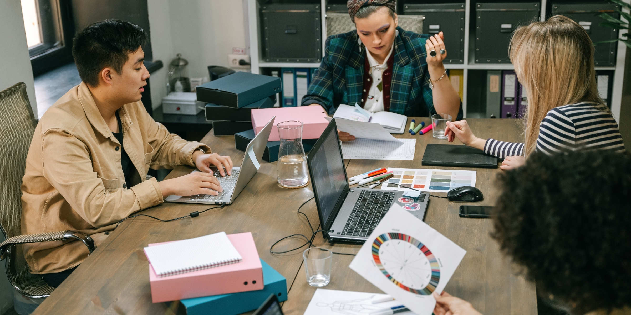 Group of diverse professionals collaborating in a modern office setting, discussing marketing strategies with laptops, color palettes, and notebooks on a wooden table, reflecting creativity and teamwork.
