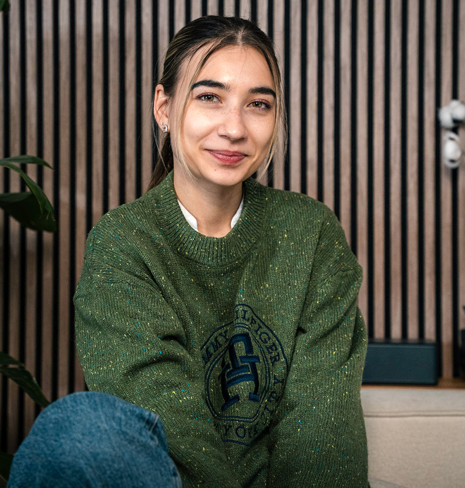 Young woman in a green sweater, smiling and seated in a modern office setting with wooden paneling, representing Kreative Angle's approachable team culture.